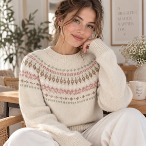 Puede incluir: Un suéter de punto de manga larga en color crema con cuello redondo y un patrón decorativo tipo Fair Isle en el pecho y los hombros en tonos suaves de verde salvia, rosa empolvado y marrón, con puños y dobladillo acanalados.