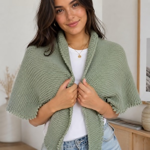 May include: A sage green knitted shawl with a collar. The shawl is worn over a white t-shirt and blue jeans. The shawl has a textured appearance and a scalloped edge.