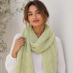 Könnte beinhalten: Ein hellgrüner Schal in offener Strickoptik mit einer zarten, luftigen Textur. Der Schal weist ein durchgehendes, sich wiederholendes Rautenmuster auf und ist aus weichem, flauschigem Garn gefertigt. Er liegt locker um den Hals über einem weißen Oberteil.