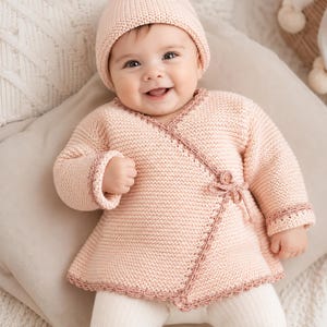 May include: A pale pink knitted baby cardigan with long sleeves, a wrap-around front closure secured by a small button and tie, and a matching knitted beanie. The garment features a textured garter stitch pattern with decorative darker pink trim.