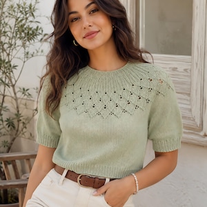 May include: A sage green short-sleeved knit jumper featuring a round neckline and a decorative lace-like diamond pattern across the upper chest and shoulders. The garment has ribbed cuffs and a ribbed hem, paired with white trousers and a brown leather belt.