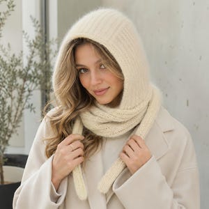 May include: A cream-coloured knitted hooded scarf featuring a ribbed texture. The accessory consists of a hood that transitions into two long, dangling scarf ends that drape down the front, crafted from a soft, fuzzy yarn material.