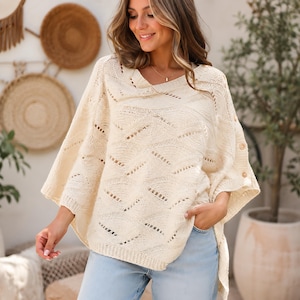 May include: A cream-colored knit poncho featuring an openwork chevron pattern, a wide neckline, and three small wooden buttons along the side seam. The garment has a loose, draped silhouette with ribbed edges at the hem and sleeve openings.
