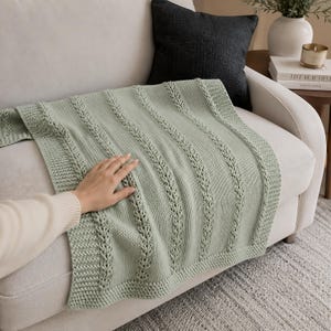 Può includere: Una coperta lavorata a maglia verde salvia con un design strutturato è drappeggiata su un divano bianco. La coperta presenta motivi a treccia verticali e un bordo a coste. Una mano è appoggiata sulla coperta. La coperta è lunga circa 152 cm.