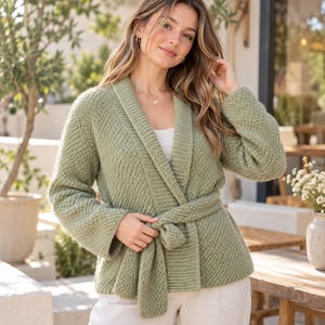 Peut inclure: Un cardigan en tricot vert sauge doté d'un col châle, de manches longues et d'une ceinture à nouer assortie à la taille. Le vêtement présente un motif tissé texturé et est porté sur un haut blanc uni avec un pantalon de couleur claire.