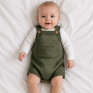 Può includere: Un bambino indossa un pagliaccetto verde oliva lavorato a maglia con bottoni in legno e una tasca frontale. Il pagliaccetto è sovrapposto a una maglietta bianca a maniche lunghe. Il bambino è sdraiato su un lenzuolo bianco e sorride.
