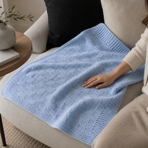 May include: A light blue knitted blanket with a textured, geometric pattern. The blanket is draped over a cream-colored armchair. The blanket's design features a series of squares and rectangles, creating a visually appealing texture. The blanket is soft and inviting.