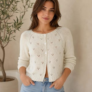Könnte beinhalten: Eine cremefarbene Strickjacke mit Rundhalsausschnitt, langen Ärmeln und einer durchgehenden Knopfleiste. Das Kleidungsstück ist mit einem durchgehenden Lochmuster an Körper und Ärmeln versehen und verfügt über gerippte Bündchen und einen Saum.