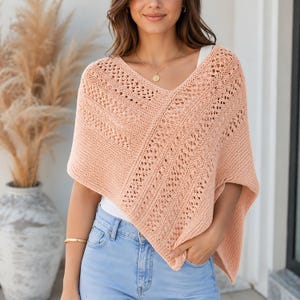 Può includere: Un poncho lavorato a maglia color pesca con scollo a V e un intricato motivo traforato. Il capo è indossato sopra un top bianco e jeans azzurri, mostrando un design a maglia testurizzata con dettagli a punto diagonale su tutto il capo.