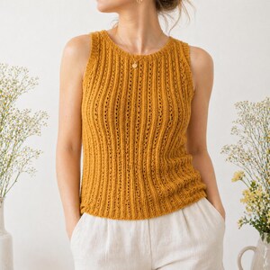 May include: Mustard yellow knitted tank top with a ribbed design. The sleeveless top has a round neckline and is made of a soft, textured yarn. The model is wearing the top with white pants. The top is a great addition to any wardrobe.