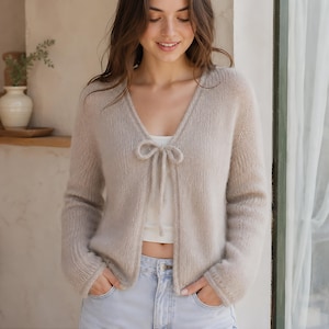 May include: A beige long-sleeved knit cardigan with a V-neckline and a delicate tie-front closure. The garment features a soft, fuzzy texture, ribbed cuffs, and a cropped length, worn over a simple white top and light blue denim jeans.