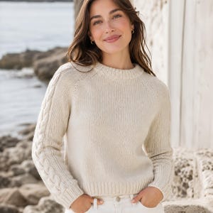 Könnte beinhalten: Ein cremefarbener Langarmpullover mit Rundhalsausschnitt und einem Zopfstrickmuster entlang der Außenseite beider Ärmel. Das Kleidungsstück ist mit einer gerippten Strickstruktur an den Ärmelbündchen, am Saum und am Kragen versehen.