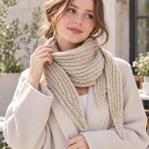 Peut inclure: Une écharpe triangulaire en tricot beige avec une texture côtelée, portée enroulée autour du cou. L'écharpe présente des extrémités pointues et une construction en grosse laine. Elle est superposée sur un haut blanc et un manteau beige.