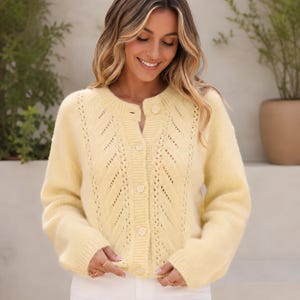 May include: A pale yellow knit cardigan featuring a round neckline, long sleeves, and a vertical openwork pattern along the front. The garment is secured with a row of matching circular buttons down the centre and has ribbed cuffs and hem.