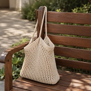 May include: A cream-coloured, open-weave tote bag with long handles, resting on a wooden bench. The bag has a textured, net-like appearance, and the handles are thick. The bench is made of dark brown wood, with a blurred background.