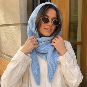 Puede incluir: Un pañuelo de punto azul claro con un patrón de textura en punto bobo. El pañuelo triangular está colocado sobre la cabeza y atado bajo la barbilla, con extremos puntiagudos que cuelgan sobre una camisa blanca de manga larga con botones.