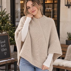 Può includere: Un poncho in maglia beige con scollo rotondo a coste e dettagli dell'orlo a coste. Il capo è realizzato in filato testurizzato ed è indossato sopra una maglia bianca a maniche lunghe e jeans blu, mostrando un design semplice e funzionale.