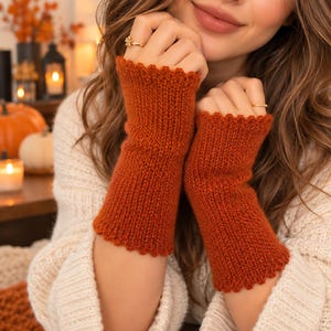 Puede incluir: Un par de guantes sin dedos de color naranja quemado con una textura de punto acanalado y un acabado de borde festoneado en la parte superior e inferior. Los guantes se usan en los antebrazos y están hechos de un hilo grueso y suave.