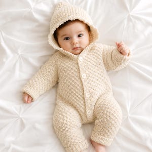 May include: A cream-colored, long-sleeved, hooded knit romper featuring a textured stitch pattern and a front button closure with four small white buttons. The garment is designed with ribbed cuffs at the wrists and ankles, providing a snug fit.