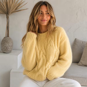 May include: A pale yellow cable-knit sweater with a crew neck. The sweater has a soft, textured appearance and is worn with white trousers. The model is smiling and has long, wavy blonde hair.