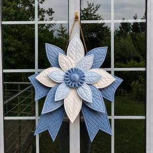 Könnte beinhalten: Eine große, gesteppte Stoffblume in Blau- und Cremetönen, die an einer Tür hängt. Die Blume hat geschichtete Blütenblätter und ein zentrales, gerüschtes Detail. Das Design ist symmetrisch und der Stoff hat eine gesteppte Textur.