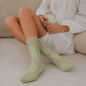 May include: A pair of sage green mid-calf socks featuring a vertical ribbed knit pattern and a decorative cable knit detail running along the side. The socks are constructed with a reinforced toe and heel section, showcasing a textured knit design.