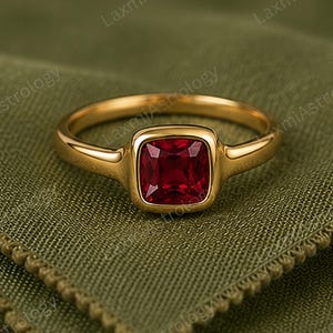 Könnte beinhalten: Ein Goldring mit einem einzelnen tiefroten Edelstein im Kissenschliff, der in einer glatten Zargenfassung sitzt. Die Ringschiene besteht aus poliertem Gold mit abgerundetem Profil und präsentiert einen leuchtend roten Stein in der Mitte.