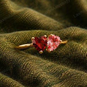 May include: A gold ring with two heart-shaped gemstones. One is a deep red, the other a light pink. The ring is set against a textured, olive-green background.