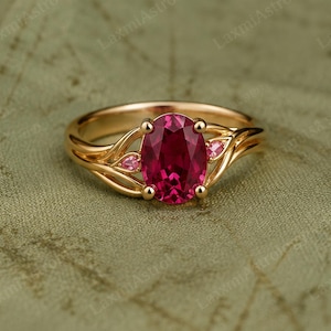 May include: A gold ring featuring a large oval-cut deep red gemstone as the centrepiece, flanked by two smaller pear-cut pink gemstones. The band has a delicate, flowing design that curves around the stones, creating an elegant, organic appearance.