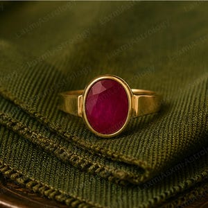 May include: A gold ring featuring an oval-cut, deep red gemstone. The ring's band is a simple, polished gold, and the gemstone is set in a gold bezel. The ring is displayed on a textured, green fabric.