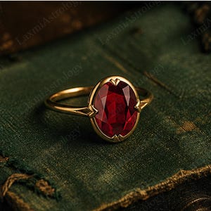 May include: A gold ring with an oval, deep red gemstone. The ring has a thin, polished gold band, and the gemstone is set in a gold bezel with decorative details. The ring is presented on a textured, dark green surface.