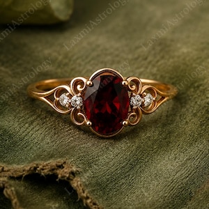 Puede incluir: Un anillo de oro con una piedra preciosa granate de color rojo intenso y corte ovalado en el centro. La banda presenta detalles de filigrana en ambos lados, acentuados por dos pequeñas piedras claras de corte redondo a cada lado del engaste.