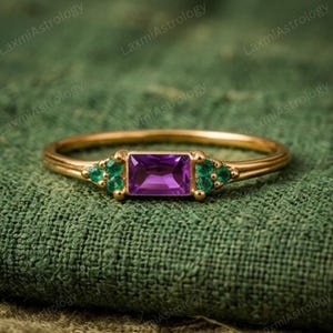 May include: A gold ring featuring a rectangular purple gemstone center, flanked by smaller green gemstones. The ring has a delicate band and is set against a textured green background. The ring is a piece of jewelry.