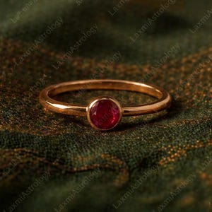 May include: A gold ring featuring a round, deep red gemstone. The ring's band is slender and polished, and the gemstone is a vibrant, striking colour. The ring is presented on a textured, dark green surface.