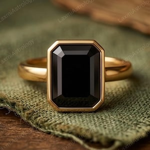 May include: A gold ring with a large, rectangular black gemstone. The gemstone is set in a gold bezel, and the band is a simple, polished gold. The ring is displayed on a green fabric.