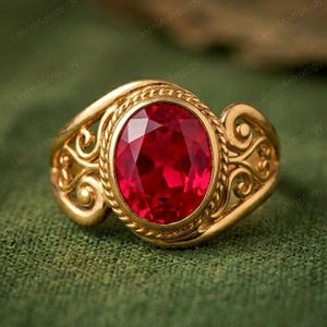 Peut inclure: Une bague en or avec une pierre précieuse rouge ovale et facettée. La bague présente un motif filigrané ornemental autour de la pierre et sur le corps. La bague est posée sur un fond vert.