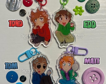 May include: Four acrylic keychains featuring cartoon characters Tord, Edd, Tom, and Matt with colored clasps. The text reads EDDSWORLD KEYCHAINS, TORD, EDD, TOM, MATT, and ART BY SCENEZZZ. Various buttons and flower-shaped charms are scattered around.
