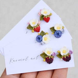 Può includere: Orecchini fatti a mano in pasta polimerica con fragole, mirtilli e more in miniatura con fiori bianchi e foglie verdi. Gli orecchini sono esposti su biglietti bianchi con la scritta "Karmel art".