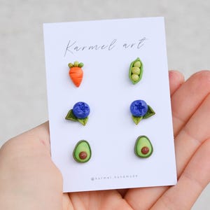 Può includere: Un set di sei orecchini a bottone fatti a mano con disegni di cibo in miniatura. Gli orecchini includono una carota, piselli, mirtilli con foglie e avocado. La carta dice "Karmel art" e "@karmel handmade".