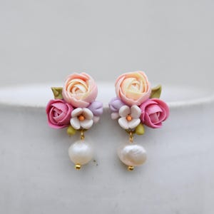 May include: A pair of stud earrings featuring clusters of sculpted pink, peach, and white flowers with green leaves. Each earring is accented with a single dangling white baroque pearl suspended from a small gold-toned metal finding.