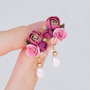 Pode incluir: Um par de brincos pendentes com cachos de rosas esculpidas em tons de rosa seco, malva e ameixa escuro, acentuados com pequenas folhas verdes. Cada brinco pende de um fio dourado com uma conta metálica e uma pérola branca.