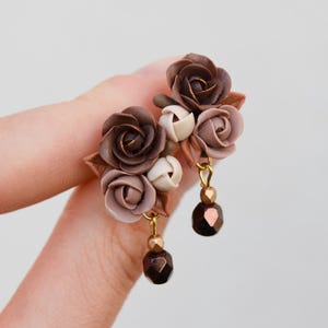 May include: A pair of stud earrings featuring clusters of sculpted roses in shades of dark brown, light brown, and cream. Each earring includes a small gold-toned metal link attached to a single dark brown faceted bead that hangs below the flowers.