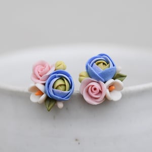 Könnte beinhalten: Ein Paar Ohrstecker mit Blumenmotiv. Jeder Ohrring zeigt eine Ansammlung von Miniaturblumen in Pastellrosa, Blau und Weiß, mit grünen Blättern. Die Ohrringe sind vor einem weichen, verschwommenen Hintergrund platziert, der die feinen Details hervorhebt.