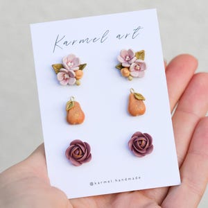 Peut inclure: Un ensemble de trois paires de boucles d'oreilles sur une carte de présentation blanche marquée Karmel art et @karmel.handmade. Les boucles présentent des grappes florales rose pâle, des poires orange avec des feuilles et des roses mauve foncé.