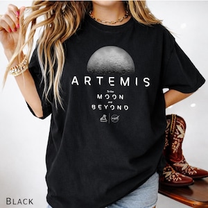 May include: Black t-shirt featuring the word "ARTEMIS" above a graphic of the moon. Below the moon graphic, the text reads "To the Moon and Beyond" with the logos of Artemis and NASA. The shirt is a comfortable fit.