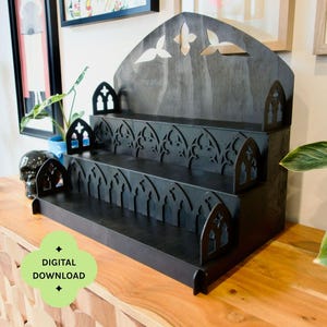 Puede incluir: Una estantería negra de tres niveles con seis estantes. La estantería tiene un diseño de arco gótico y recortes decorativos. La parte superior de la estantería presenta formas recortadas. El texto "DESCARGA DIGITAL" y "CONTIENE SEIS ESTANTES" son visibles.