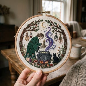 May include: Embroidered artwork in a wooden hoop shows a witch in a green dress adding herbs to a bubbling cauldron. A black cat sits nearby. The scene includes bottles, mushrooms, and a crescent moon, all in a whimsical style.