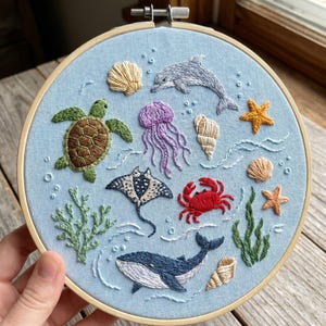 Pode incluir: Um bastidor de bordado circular de madeira contendo tecido azul claro com vida marinha bordada à mão, incluindo uma tartaruga marinha verde, uma água-viva roxa, um caranguejo vermelho, uma baleia azul, um golfinho cinza, uma arraia, várias conchas, estrelas-do-mar e algas verdes.