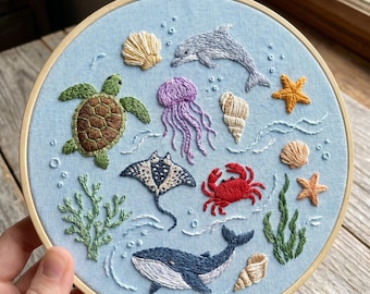 May include: A circular wooden embroidery hoop containing light blue fabric with hand-stitched marine life including a green sea turtle, a purple jellyfish, a red crab, a blue whale, a grey dolphin, a stingray, various seashells, starfish, and green seaweed.