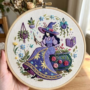 Op de afbeelding: Geborduurde hoepelkunst met een heks in een paarse jurk en hoed, die een toverstaf vasthoudt. Het ontwerp omvat een ketel, bloemen, hangplanten en een boek, allemaal in paarse, groene en gele tinten. Het kunstwerk is gevat in een houten hoepel.